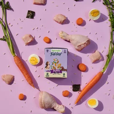 Naturediet Feel Good Puppy food with visible ingredients including chicken pieces, carrots, rice grains, hard-boiled eggs and seaweed arranged around the product packaging.
