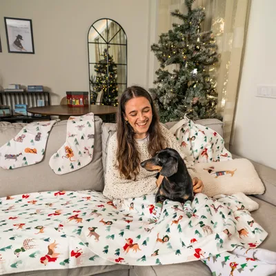Manta, almofada e meias de Natal com padrão de cães e árvores, expostas num sofá ao lado de uma árvore de Natal decorada. Cão pequeno sentado no colo de pessoa.