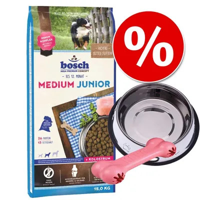 Confezione Bosch Medium Junior 15 kg, ciotola in metallo, osso rosa in gomma, simbolo percentuale rosso per offerta. Testo visibile: 'BIS 12. MONAT', 'KOLOSTRUM', '26 PROTEIN', '13 FETTGEHALT'.