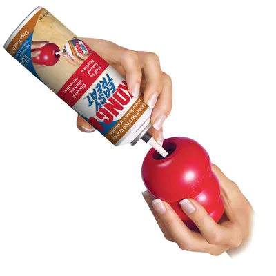 Hand squeezing treat paste from a can into a red toy, demonstrating how to fill the toy with the product for use.
