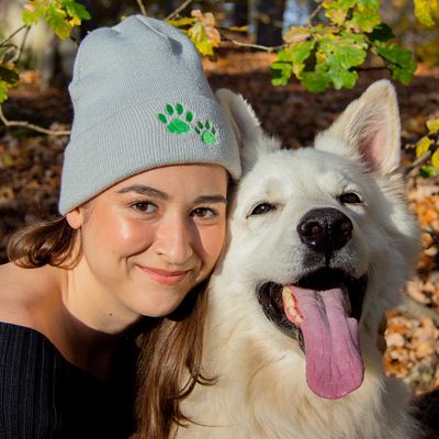 Fehér kutya nyitott szájjal és kilógó nyelvvel, mellette egy személy szürke sapkában zöld mancsnyomattal.