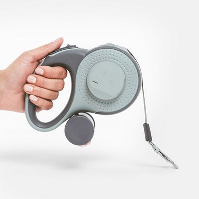 Mão segura uma trela cinzenta para cães com pega ergonómica, dispensador integrado e fita extensível com clip metálico.