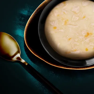 Nahaufnahme einer Schüssel mit heller Suppe, sichtbar sind Stücke von Huhn und Gemüse. Daneben ein goldener Löffel auf dunklem Hintergrund.