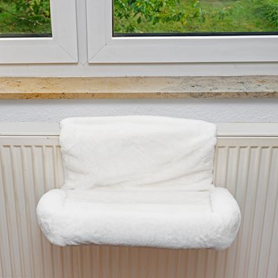 Weiße Heizkörperliege mit weichem Bezug, montiert an einem Heizkörper unter einem Fenster.