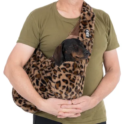 Petit chien noir dans un sac de transport en tissu à motif léopard, porté en bandoulière par une personne. Marque visible : TIAKI.