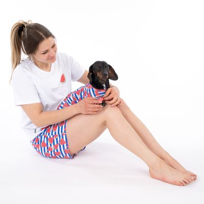Frau in weissem T-Shirt und Shorts mit Wassermelonenmuster hält einen kleinen Hund, der ein passendes Outfit mit Wassermelonenmuster trägt.