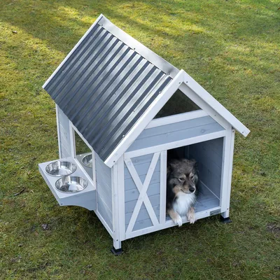 Gråt hundehus i træ med sort metaltag, to foderskåle i rustfrit stål på siden og en hund indeni. Står på en grøn græsplæne.