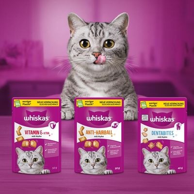 Whiskas Snacks s kuřetem: Vitamin E-Xtra 50g, Anti-Hairball 50g, Dentabites 40g. Méně plastu, stejná chuť. Kočka v pozadí.