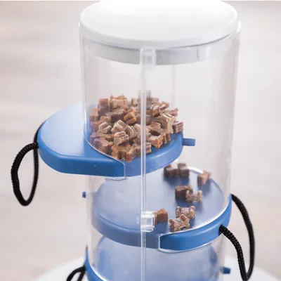 Transparante voederdispenser met blauwe inzet, gevuld met kleine stervormige snacks. Twee zichtbare niveaus, verbonden met zwarte koorden.