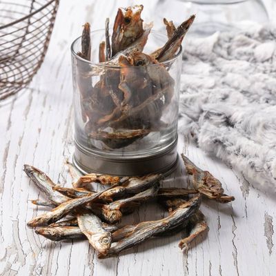 Dried whole fish treats displayed in a glass and scattered on a wooden surface, showing the product’s size and texture.