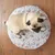 Coussin rond pour chien avec housse à motifs de chiens et d’os, un carlin allongé dessus, posé sur un sol en bois. Matière textile visible.