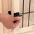 Fermeture en métal noir sur porte en bois et grille, main actionnant le loquet. Matériaux visibles : bois clair et métal noir.