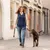 Frau führt einen braunen Hund an einer Leine mit Geschirr durch eine Stadtstrasse. Hund trägt ein schwarzes Geschirr.