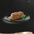 Portion of wet pet food on a black plate with a sprig of parsley garnish, shown against a dark background with water droplets. No visible brand or text.