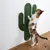 Chat tigré debout sur un griffoir mural en forme de cactus, fixé au mur, matériau corde verte visible. Sol en bois clair.