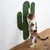 Chat utilisant un griffoir mural en forme de cactus, recouvert de corde verte, fixé sur un mur blanc au-dessus d'un sol en bois clair. Aucun texte ou marque visible.