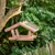 Casita para pájaros colgante de madera con techo verde y zona de comida, llena de alimento para aves, fijada a un árbol.