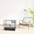 Cage pour petits animaux en métal et plastique noir, avec plateforme rose, maisonnette verte et abreuvoir, posée sur un sol en bois clair dans un intérieur moderne.