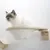 Wall-mounted cat lounger made of wood with white woven fabric. Cat sitting on it, looking forward.