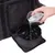 Main versant de l’eau d’un verre dans une gamelle pliante noire en tissu, posée devant un sac noir à poche zippée visible en arrière-plan.