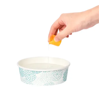 En hand häller vätska från en gul mätkopp i en vit skål med blått blommönster.