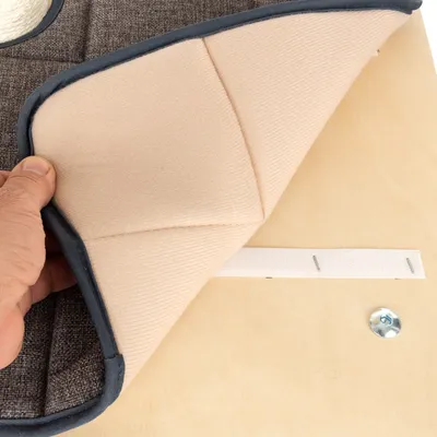 Hand hebt eine beige gepolsterte Matte mit Klettverschluss von einer Holzplatte ab. Sichtbar sind Schraube und grauer Stoff darunter.