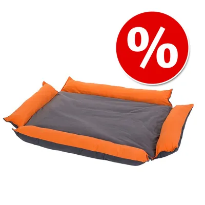 Cama rectangular para mascotas en tela gris con bordes naranjas. Círculo rojo con símbolo de porcentaje blanco, indicando oferta o descuento visible.