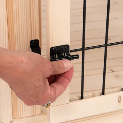 Fermeture en métal noir sur porte en bois et grille, main actionnant le loquet. Matériaux visibles : bois clair et métal noir.