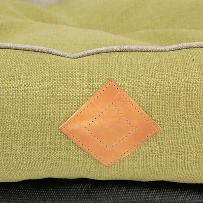 Nærbillede af grøn tekstil med synlig brun læderdetalje i kvadratisk form, kontrastsyning og beige kant. Materialer: stof og læder.