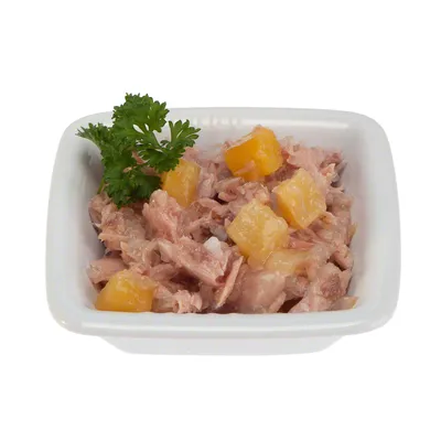 Влажный корм для животных в белой миске: кусочки мяса с жёлтыми кубиками овощей, украшено веточкой петрушки. Видны текстура и состав продукта.