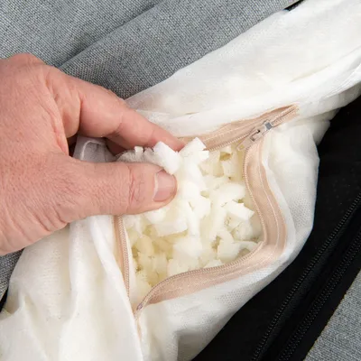 Mano che mostra imbottitura interna in schiuma a cubetti all'interno di una fodera con cerniera, su tessuto grigio. Materiale visibile: schiuma, tessuto.