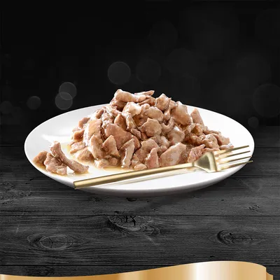 Кусочки влажного корма на белой тарелке с золотистой вилкой, корм в соусе или желе, без видимых брендов или текста.