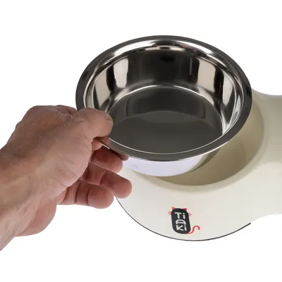 Hand holding a removable stainless steel bowl over a white food bowl with Tiaki logo.