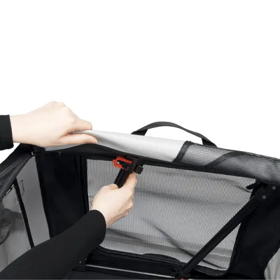 En hand visar hur den röda låsmekanismen på en svart transportbur öppnas. Nätmaterial och bärhandtag synliga.