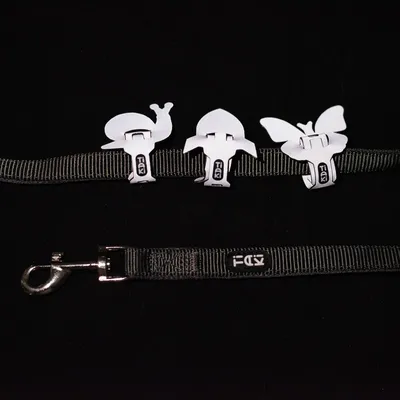 Cinta negra con tres clips blancos en forma de animales: caracol, pez y mariposa. Gancho metálico visible al final de la cinta.