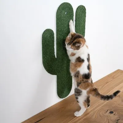 Arranhador de parede em forma de cato, feito de corda verde, com um gato a arranhar. Produto montado numa parede branca junto a uma superfície de madeira.