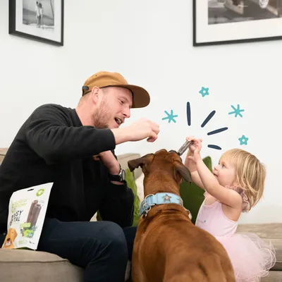 Chien avec un collier bleu recevant une friandise de l'emballage 'Busy Treat' donnée par une personne. L'emballage montre une friandise pour chien avec des accents verts.