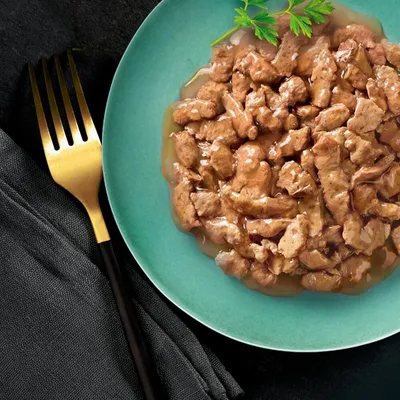 Assiette verte contenant des morceaux de nourriture humide en sauce, garnie d'une branche de persil. Fourchette dorée posée à côté sur une serviette noire.