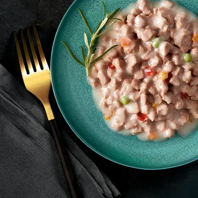 Close-up of a plate with meat-based wet food, vegetables such as peas and bell pepper in sauce, garnished with a sprig of rosemary. Golden fork and black cloth beside.