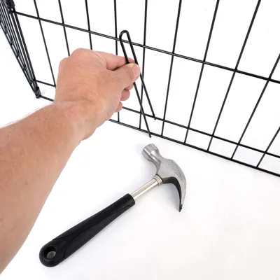 Main tenant une tige métallique devant un enclos grillagé noir, marteau à manche noir posé au sol à côté.