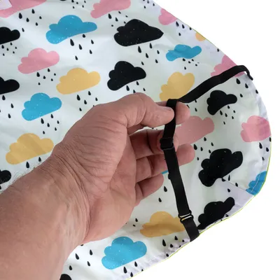 Main tenant un élastique réglable fixé à un tissu imperméable décoré de nuages colorés et gouttes de pluie, montrant le système d’attache du produit.