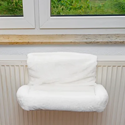 Weiße Heizkörperliege mit weichem Bezug, montiert an einem Heizkörper unter einem Fenster.
