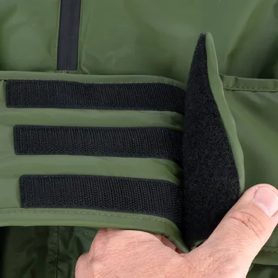 Gros plan sur une fermeture à scratch noire sur un manteau vert, tenue par une main. Matière extérieure lisse, intérieur en tissu doux visible.