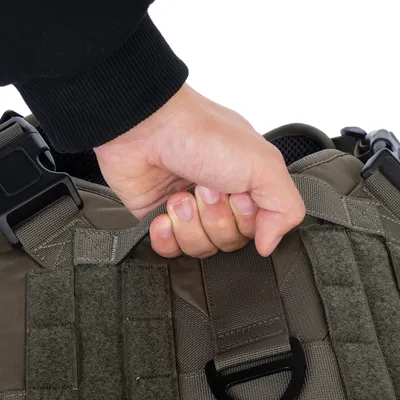 Hand greift an einem stabilen Griff eines olivgrünen Hundegeschirrs mit Klettflächen und Schnallen.