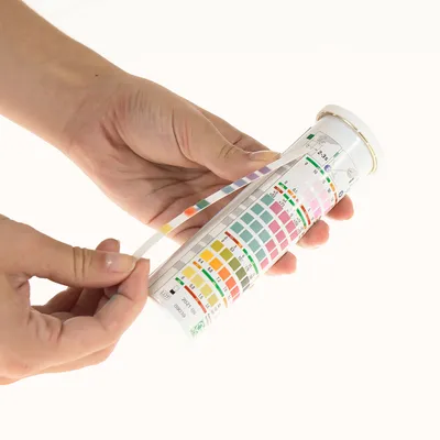 Ręka trzyma pasek testowy i skalę barwną do analizy parametrów wody. Skala pokazuje różne kolory dla pH, azotynów, azotanów i innych parametrów.