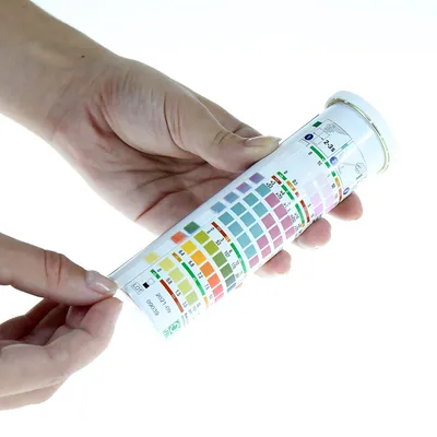 Ręka trzyma pojemnik z paskami testowymi z kolorową skalą do analizy parametrów wody. Skala pokazuje pH, azotany i chlor.