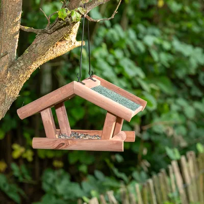 Casita para pájaros colgante de madera con techo verde y zona de comida, llena de alimento para aves, fijada a un árbol.