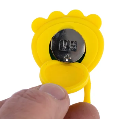 Gelbes Batteriefach mit geöffneter Abdeckung, zeigt eine eingelegte Knopfzellenbatterie. Hand hält das Fach zur Demonstration.