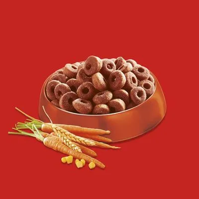 Ronde brokjes in een metalen voerbak, met zichtbare wortels, tarwe en maïskorrels ernaast. Geen merknaam of tekst zichtbaar.