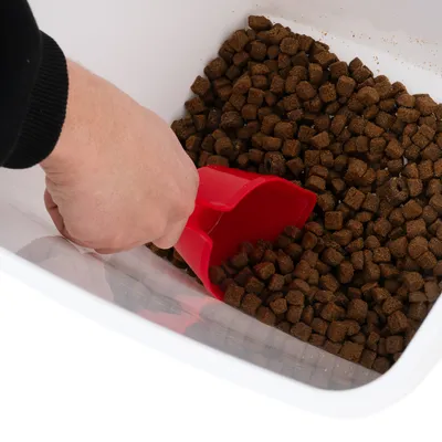 Hand with red scoop taking brown dry food pieces from a white container.